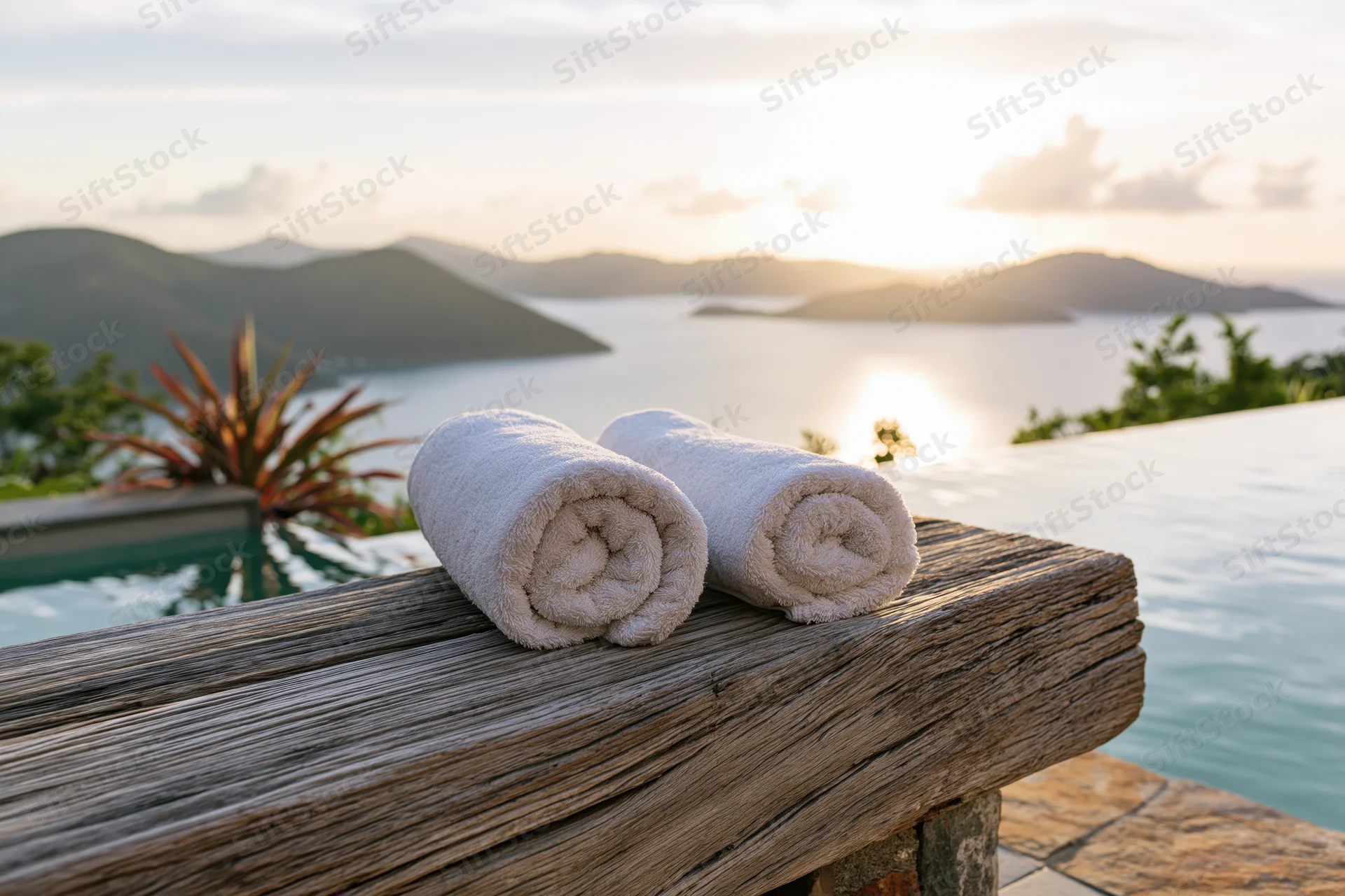 Two rolled white towels on a weathered teak bench beside an infinity pool overlooking ocean and mountains at sunset