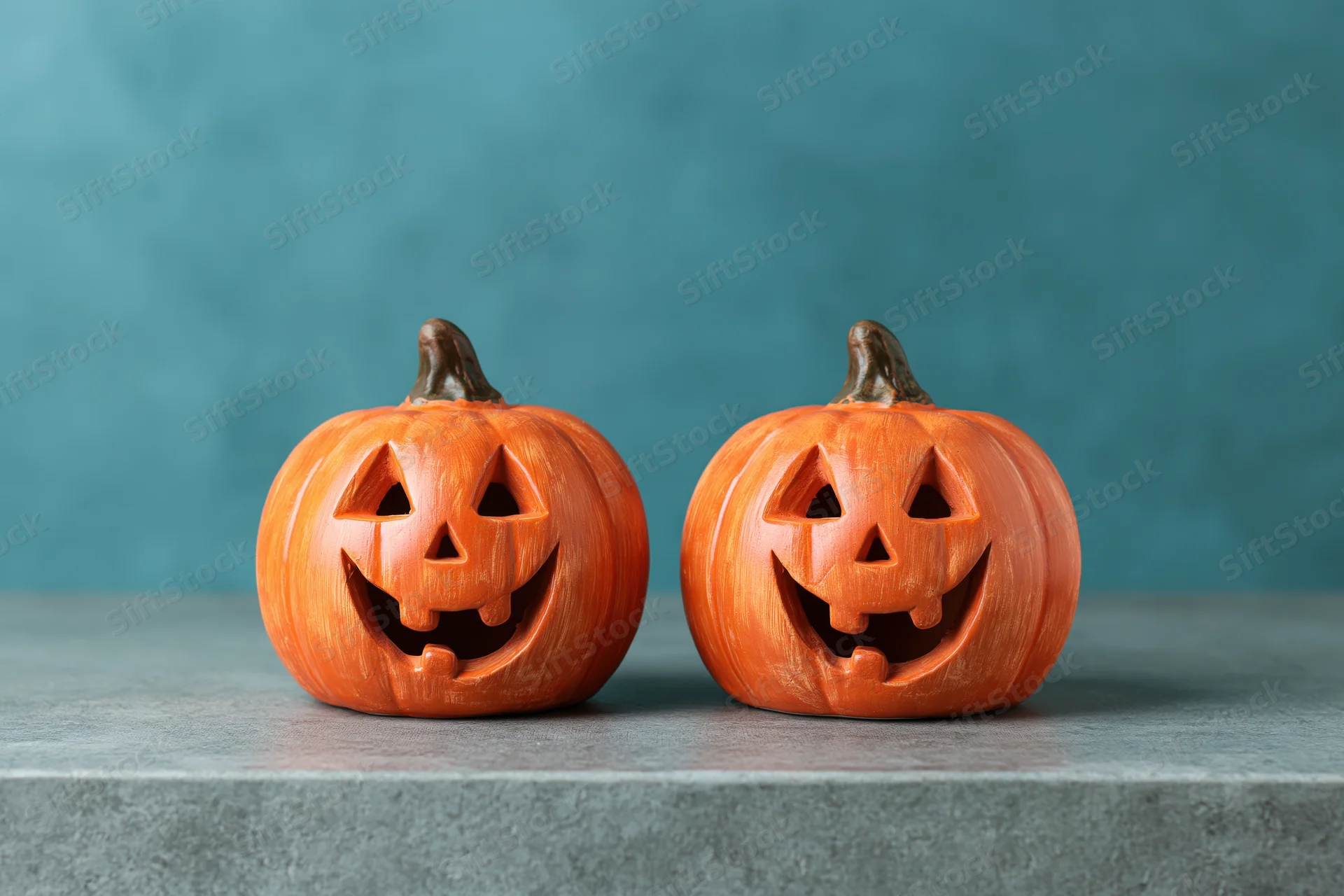 Two small ceramic jack-o-lantern figurines with carved smiling faces on a gray surface against a teal background
