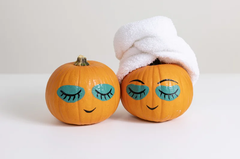 Two small orange pumpkins with painted closed eyes and smiles, one wearing a white towel, on a white surface