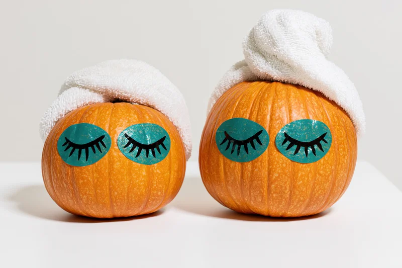 Two small orange pumpkins with painted closed eyes and white towels on a white surface