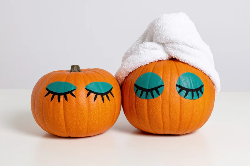 Two small orange pumpkins with painted closed eyes, one wearing a white towel, on a white surface