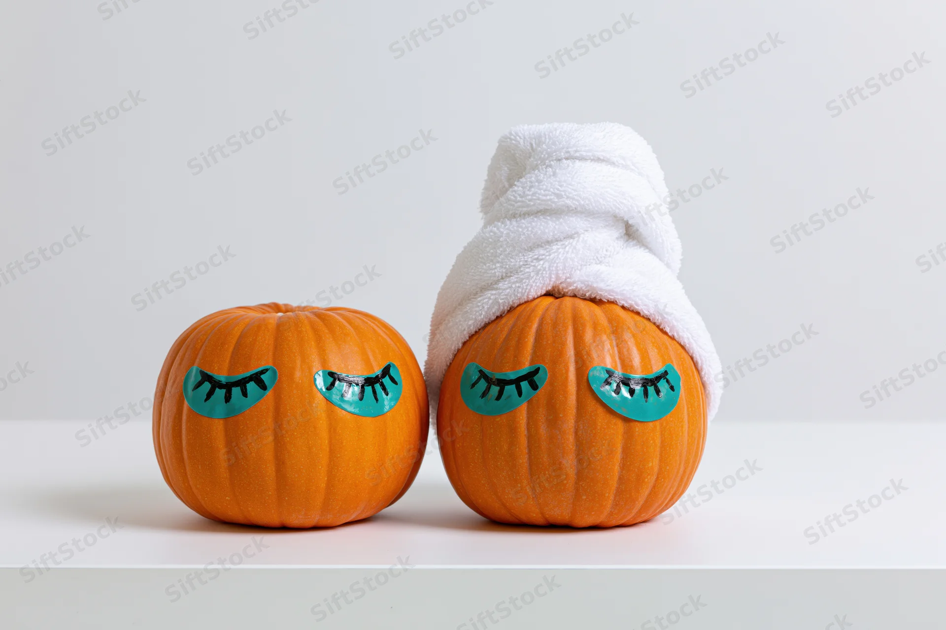 Two small orange pumpkins with painted closed eyes, one wrapped in a white towel, on a white surface