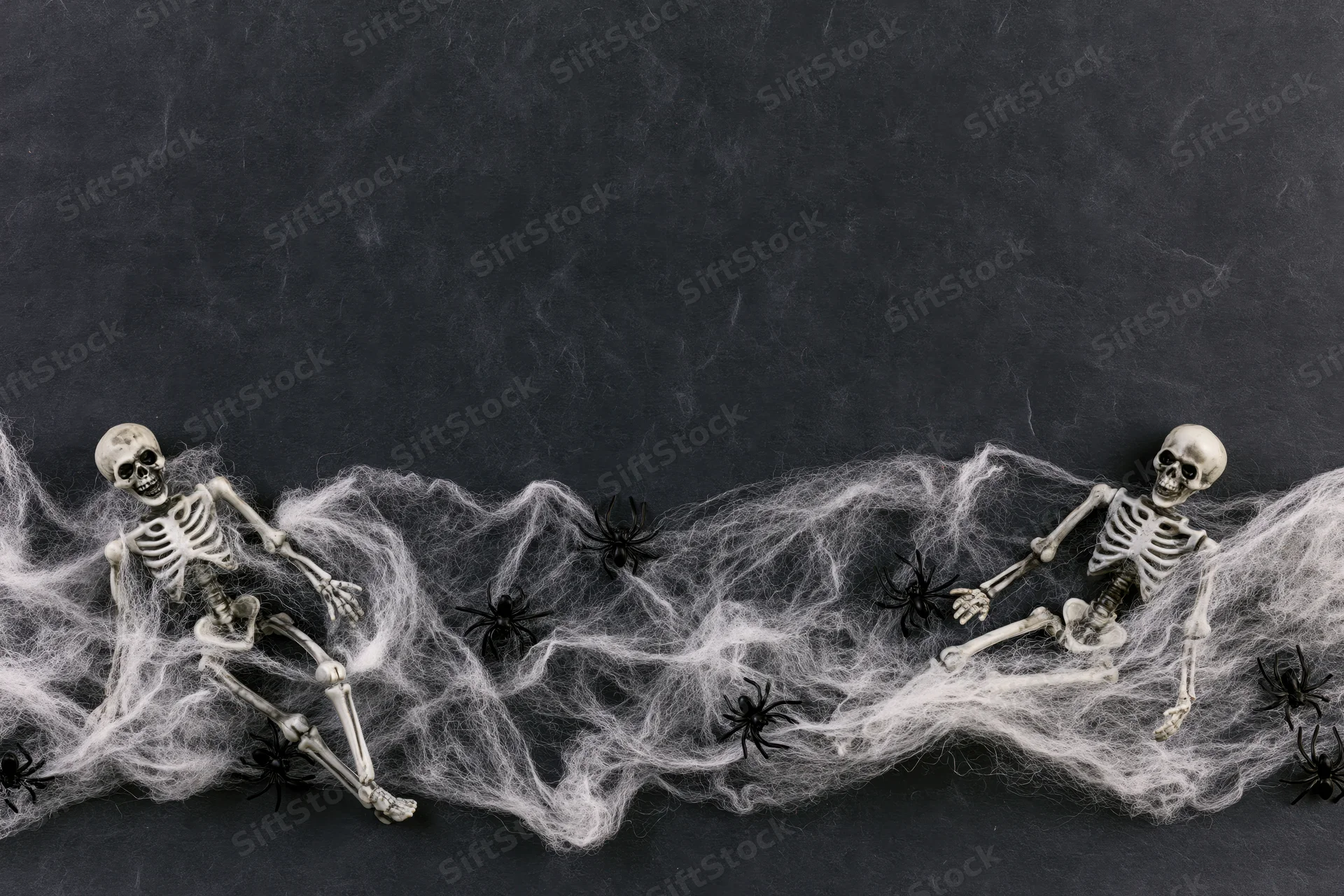 Two small plastic skeleton figurines tangled in white cobweb with black spiders on dark surface