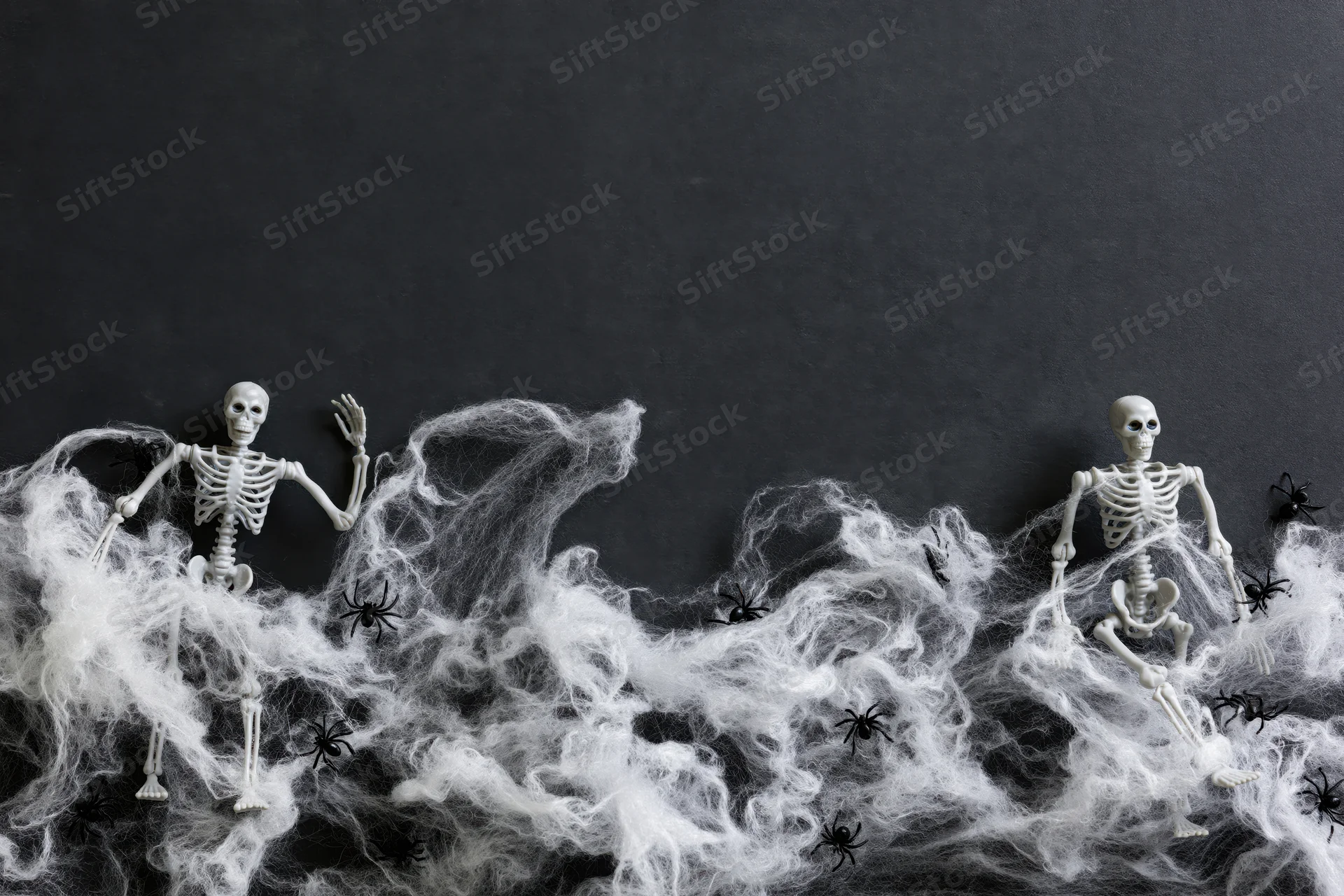 Two small plastic skeleton figurines tangled in white cobweb with black spiders on dark surface