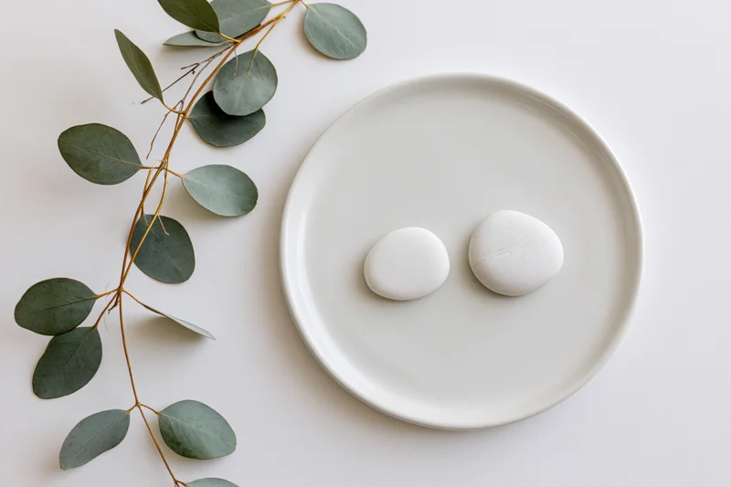Two smooth white stones on a round white ceramic plate beside a eucalyptus branch on a plain white surface