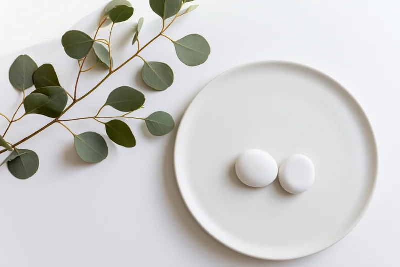 Two smooth white stones on a round white ceramic plate beside a eucalyptus branch on a white surface