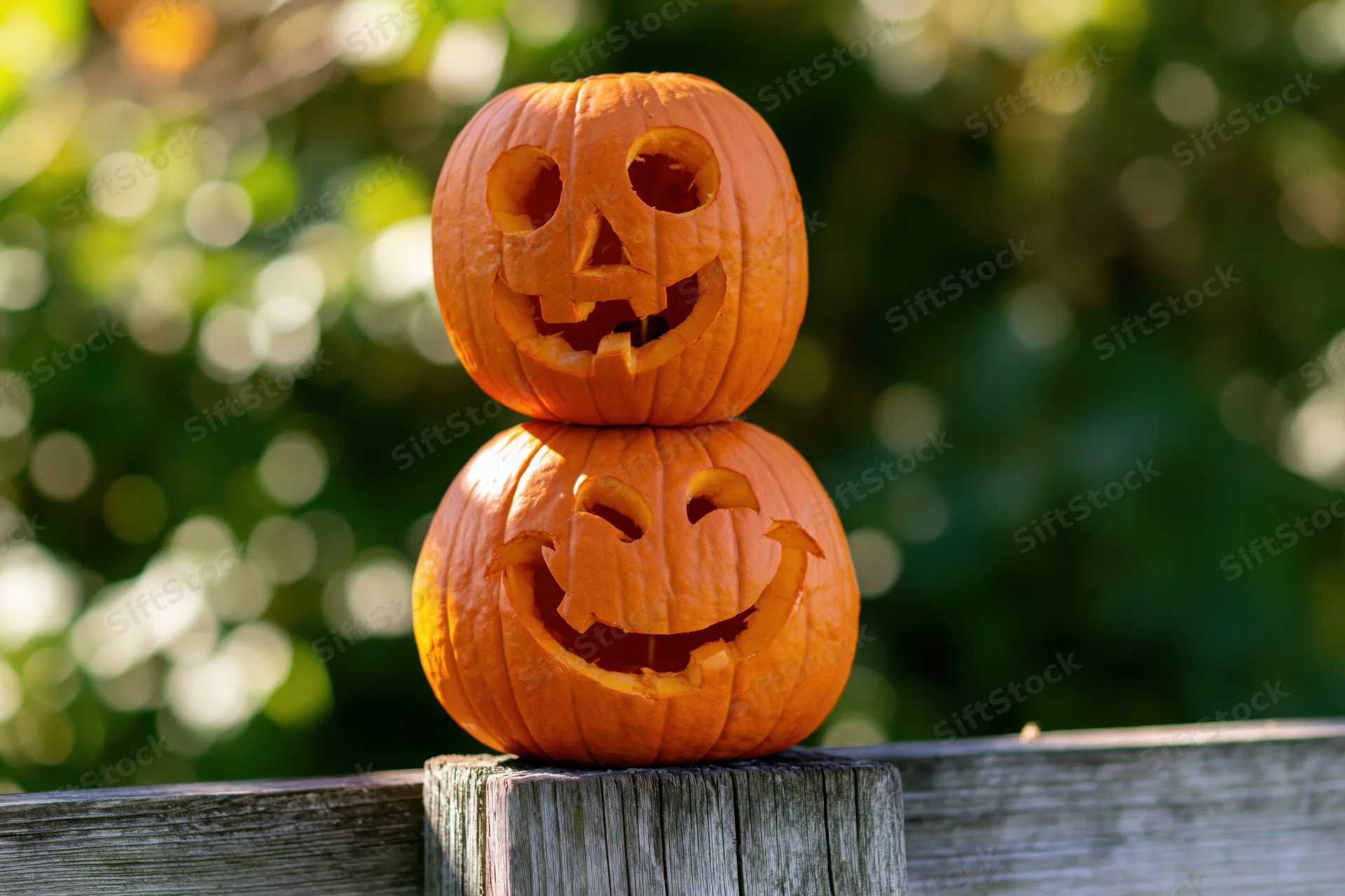 Two carved pumpkins stacked on a wooden fence outdoors