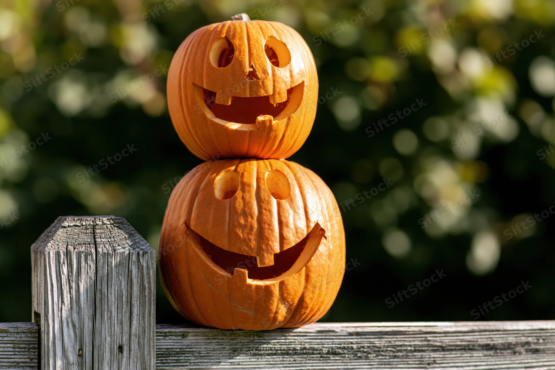 Two carved pumpkins stacked on a wooden fence outdoors
