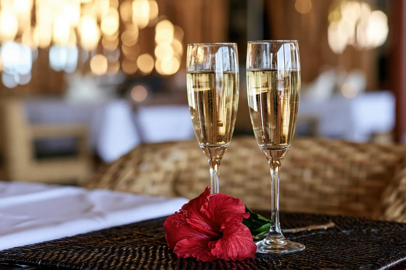 Two champagne flutes filled with sparkling wine on a table with a red hibiscus flower and woven placemat in a restaurant setting