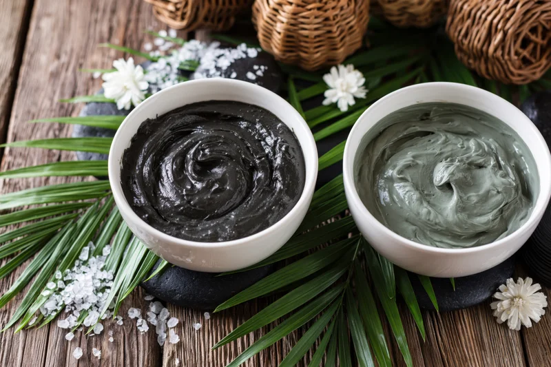 Two white ceramic bowls with black and gray facial masks on a wooden table with palm leaves and flowers
