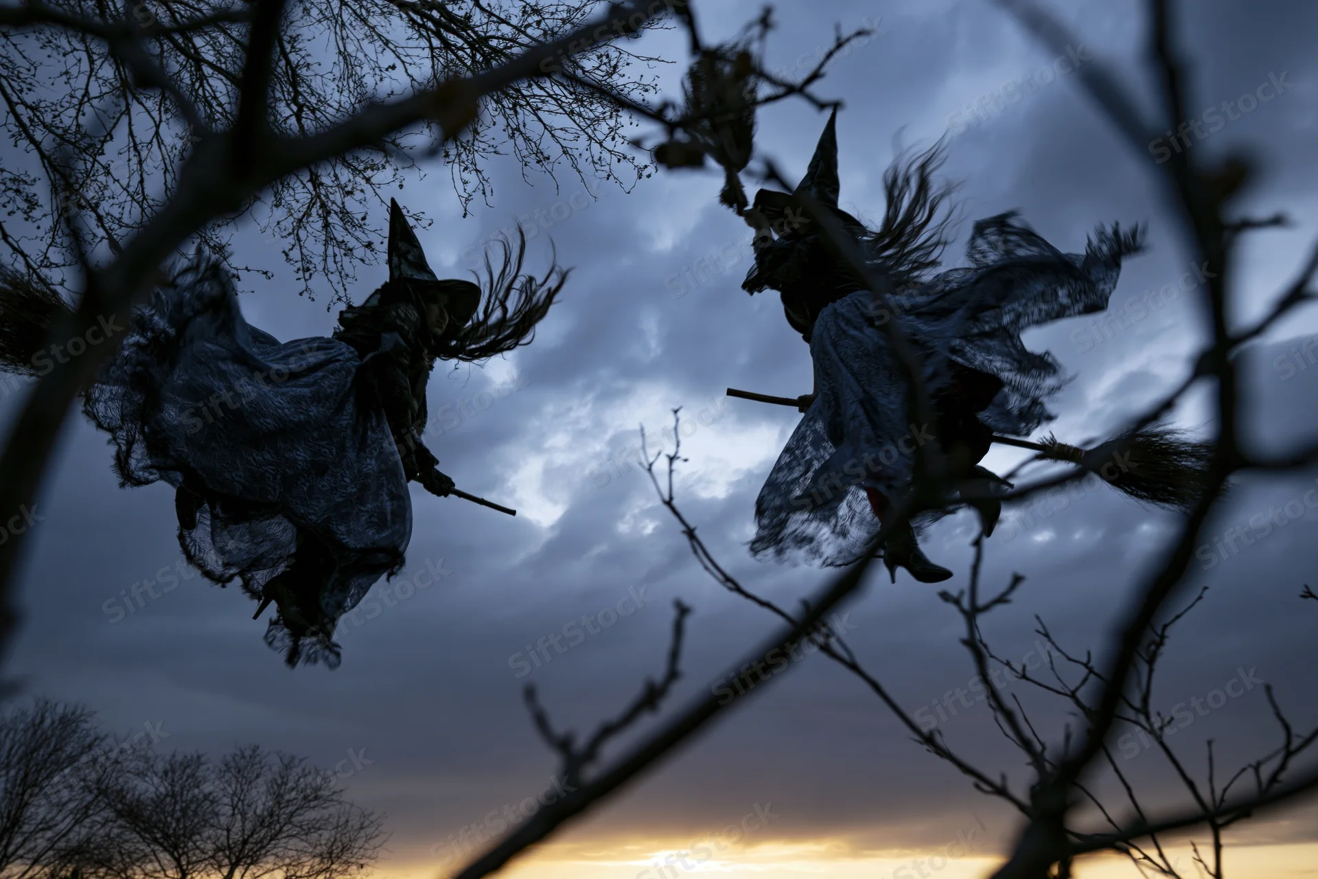 Two women in witch costumes flying on broomsticks silhouetted against a cloudy twilight sky