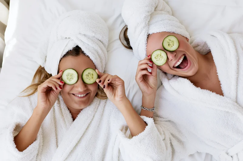 Two women lying on white bedding with cucumber slices over their eyes, wearing bathrobes and towels