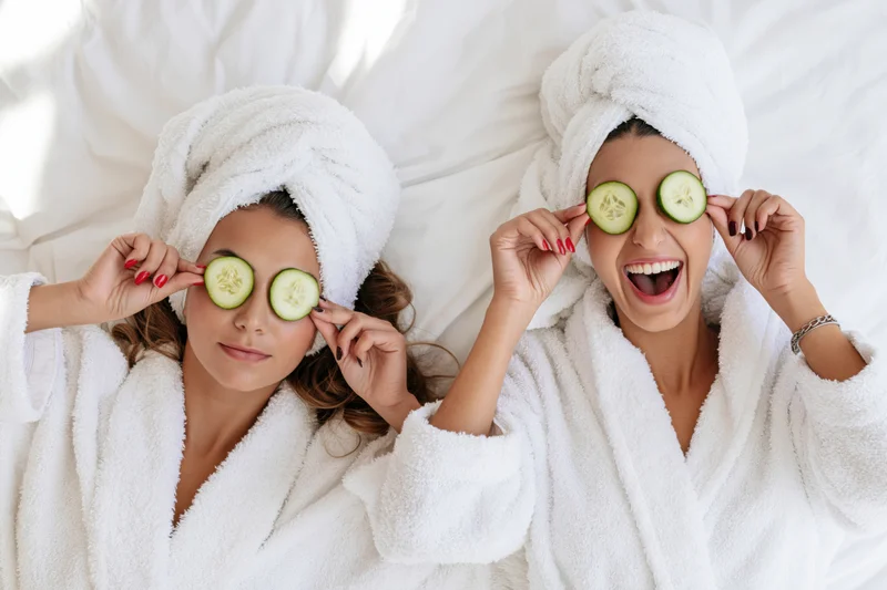 Two women lying on white bedding with cucumbers over their eyes wearing bathrobes and towels