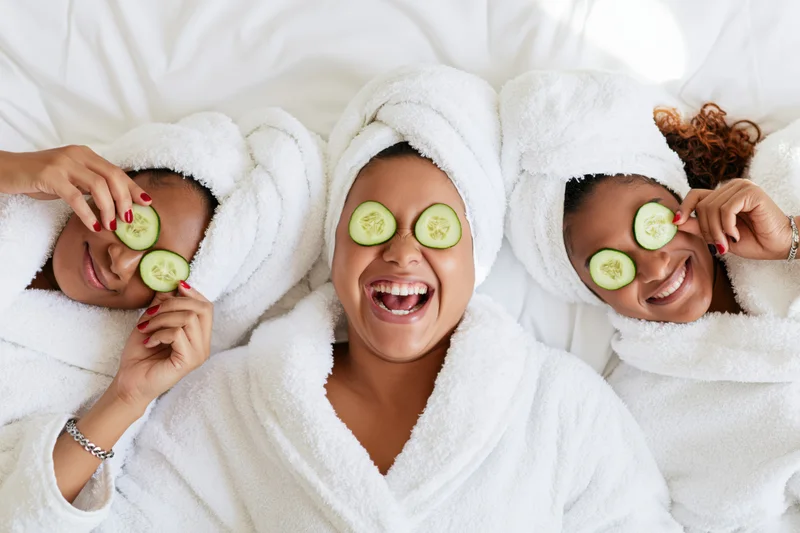 Three women in white robes with cucumbers on eyes laughing on white bedding
