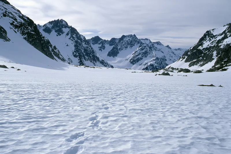 Snow-covered alpine valley floor with footprints leading toward jagged mountain peaks under overcast sky