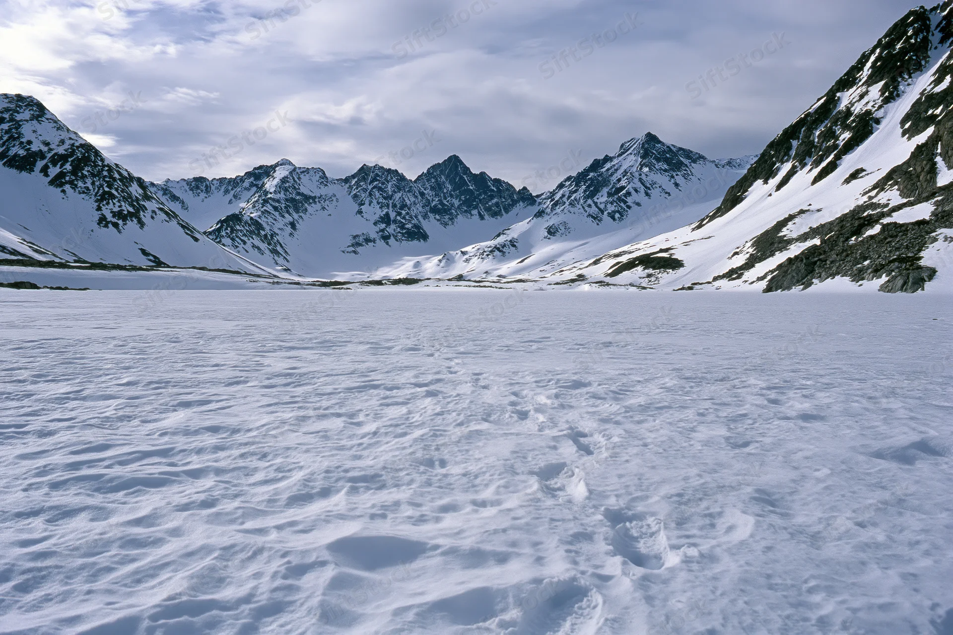 Snow-covered alpine valley floor with footprints leading toward jagged mountain peaks under cloudy sky