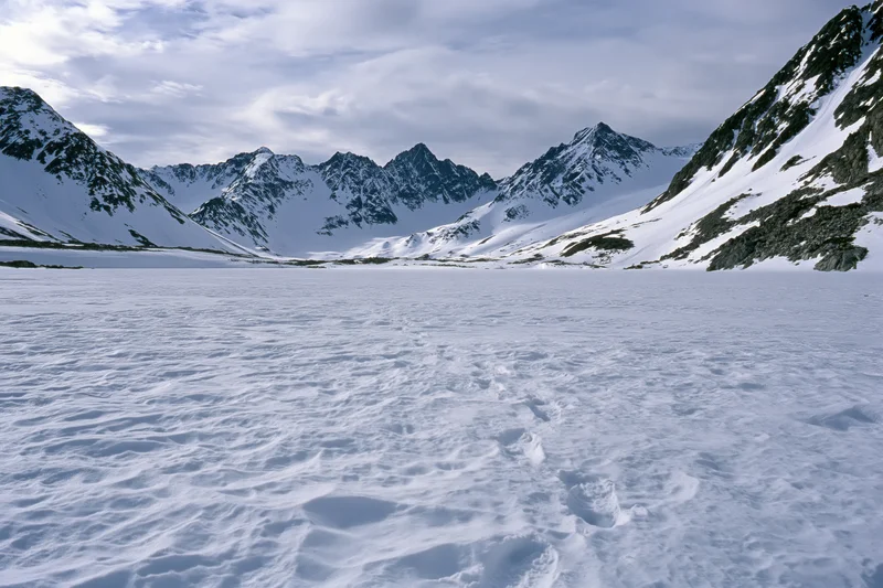Snow-covered alpine valley floor with footprints leading toward jagged mountain peaks under cloudy sky