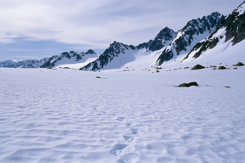 Snow-covered alpine valley floor with footprints leading toward jagged peaks under a partly cloudy sky