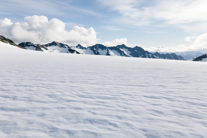 Vast snow-covered glacier plateau stretching toward jagged mountain peaks under a partly cloudy sky