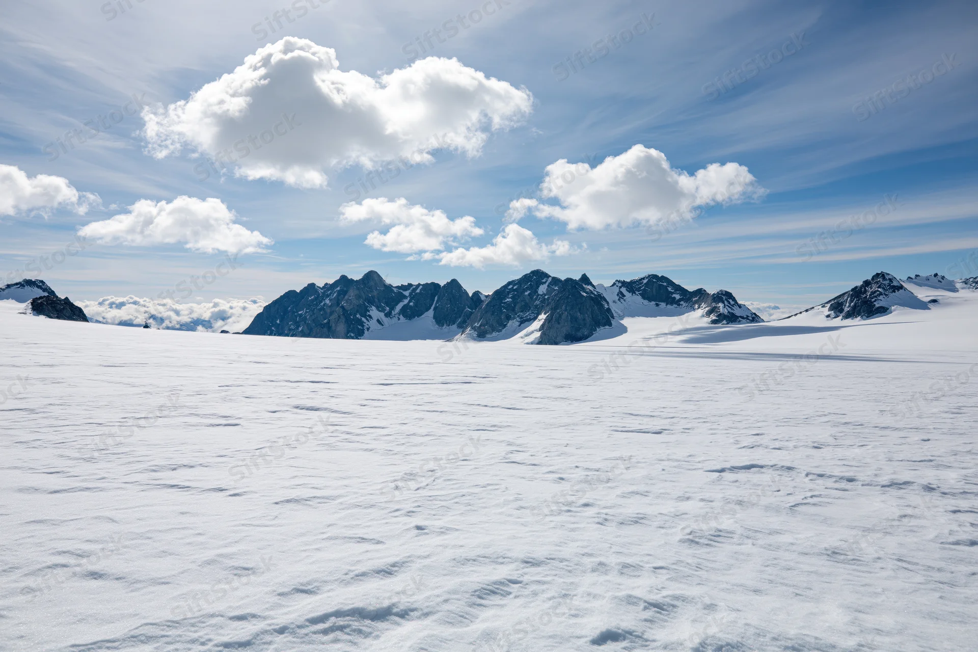 Vast snow-covered glacier plateau stretching toward jagged mountain peaks under a partly cloudy sky