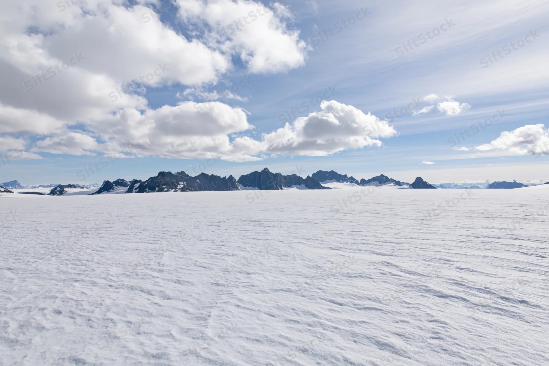 Vast snow-covered glacier plateau stretching toward jagged mountain peaks under a partly cloudy sky