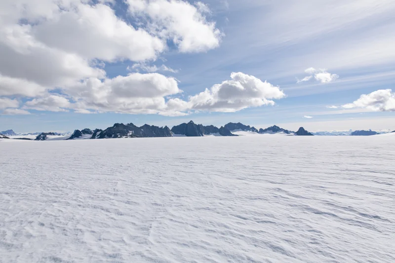 Vast snow-covered glacier plateau stretching toward jagged mountain peaks under a partly cloudy sky