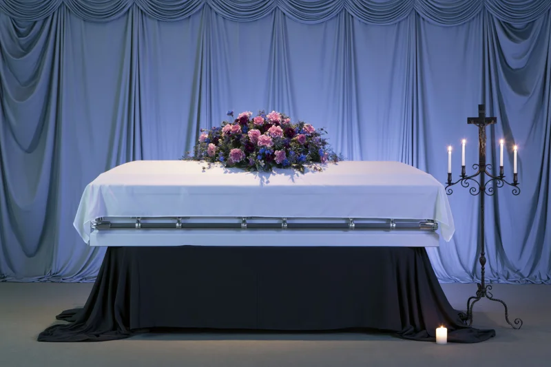 White coffin on draped catafalque with floral arrangement and lit candles in funeral chapel