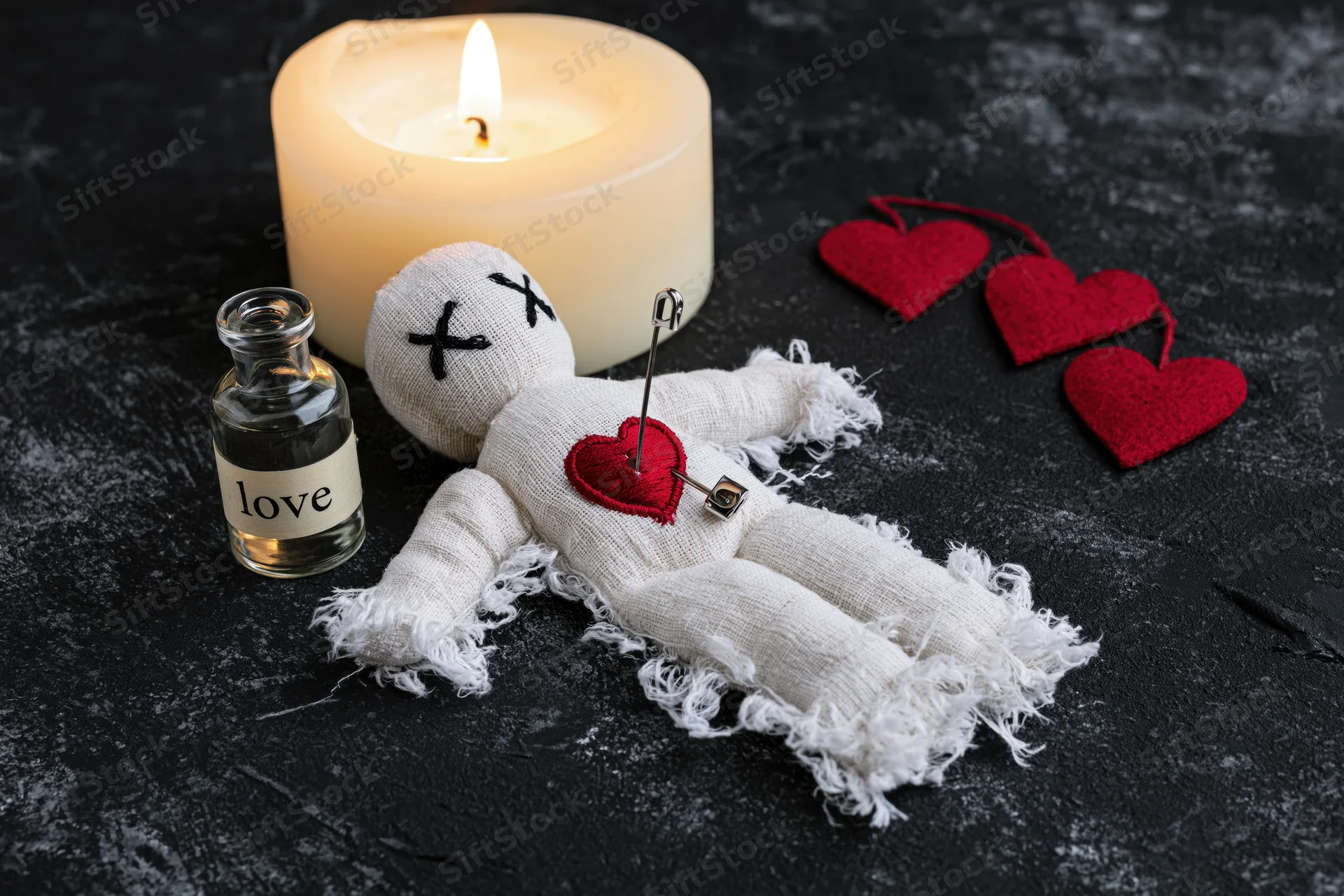 White fabric voodoo doll with red heart and pin lying on dark textured surface beside lit candle and bottle labeled love