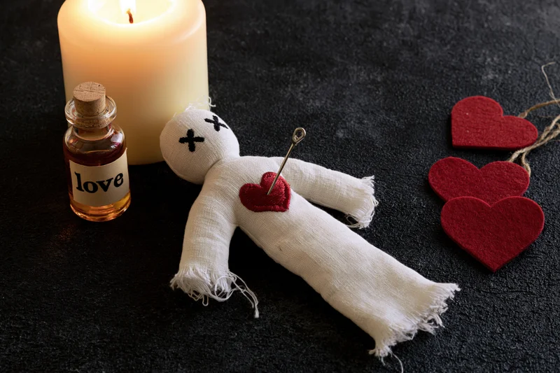 White fabric voodoo doll with needle in red heart lying on dark surface beside candle and bottle labeled love