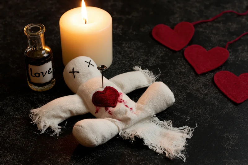 White fabric voodoo doll with red heart and needle lying on dark surface beside lit candle and bottle labeled love