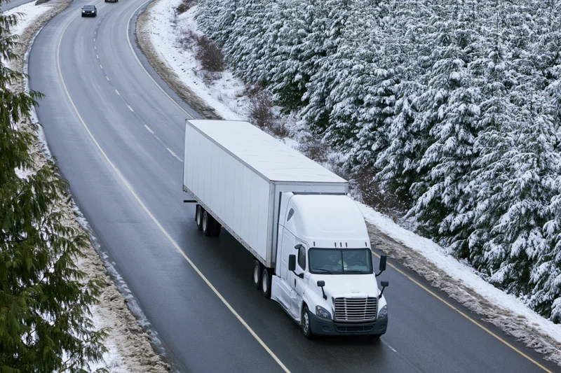 White semi-truck with dry van trailer driving on wet winter highway surrounded by snow-covered evergreen trees