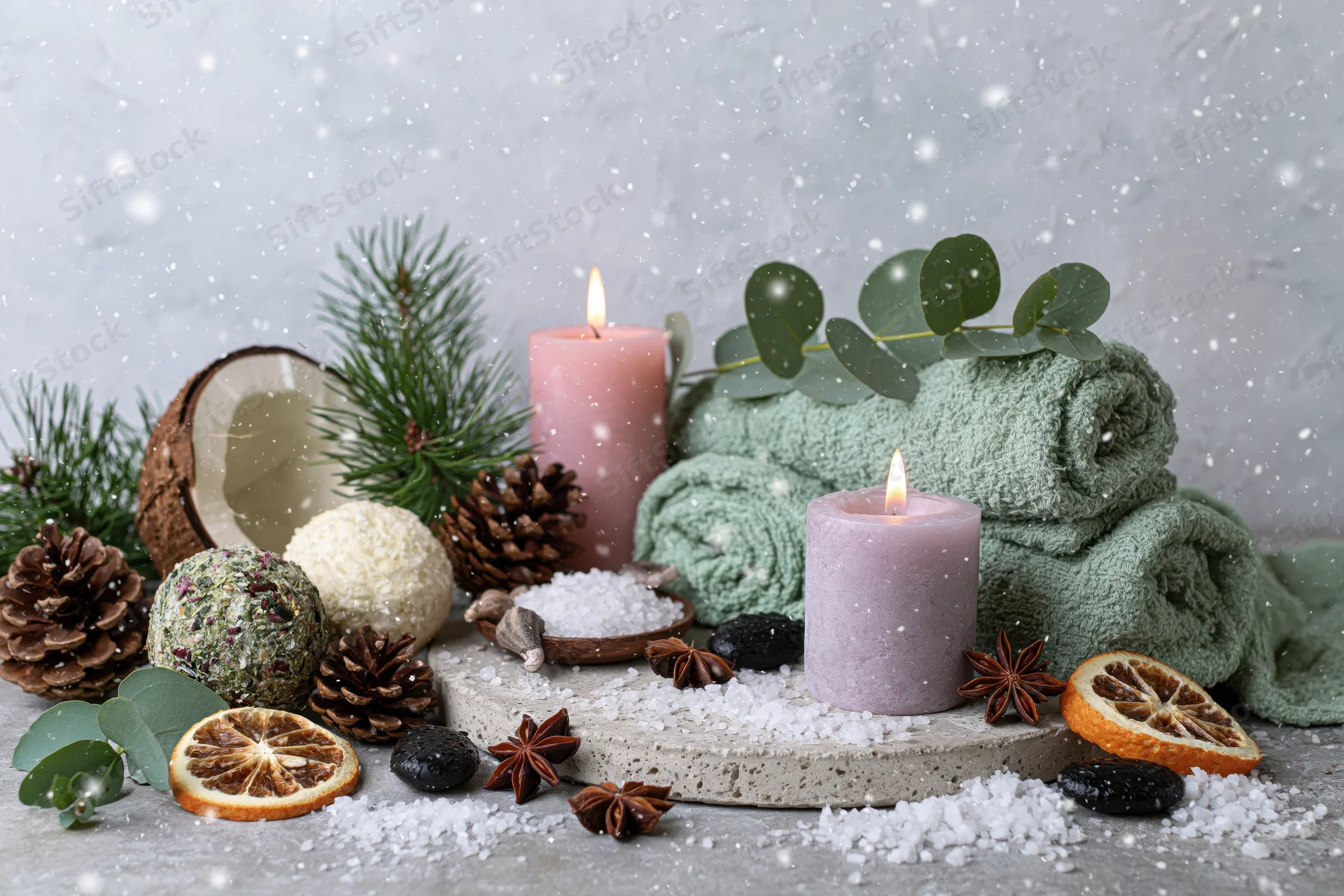 Winter spa flat lay with lit candles, towels, pine, eucalyptus, and bath salts on grey concrete surface with falling snow