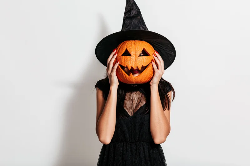 Woman in black witch costume holding carved pumpkin over face against white background