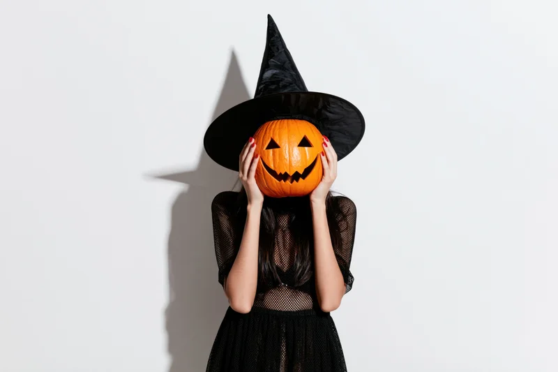 Woman in black witch costume holding carved pumpkin over face against white background