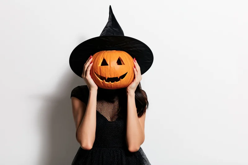 Woman in black witch costume holding carved pumpkin over face against white background