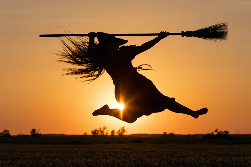Witch silhouette flying on broomstick midair over field at sunset