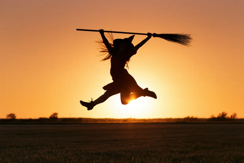 Woman in witch costume jumping with broomstick against sunset over field