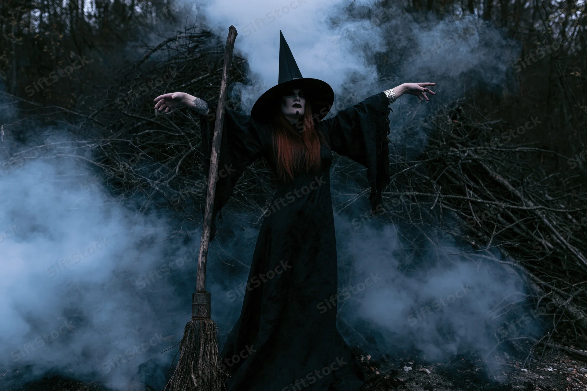 Witch in black costume with pointed hat and broom standing in smoky forest with arms outstretched