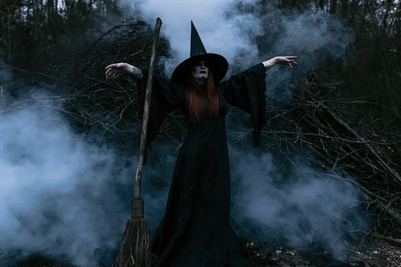 Witch in black costume with pointed hat and broom standing in smoky forest with arms outstretched