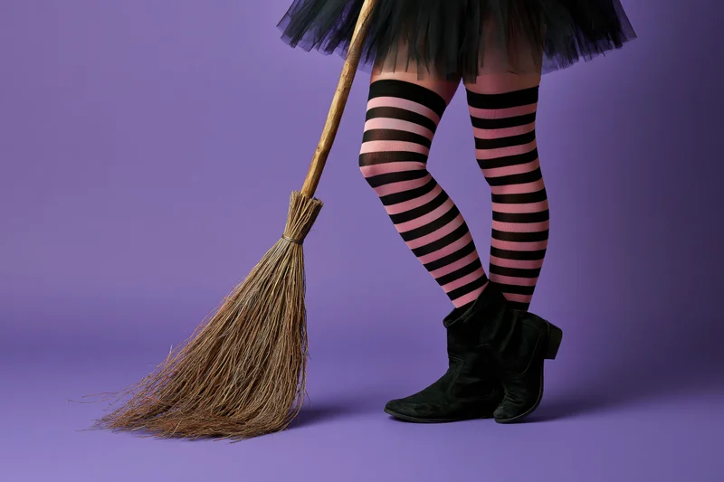Woman's legs in striped stockings and black boots holding a broom against purple background