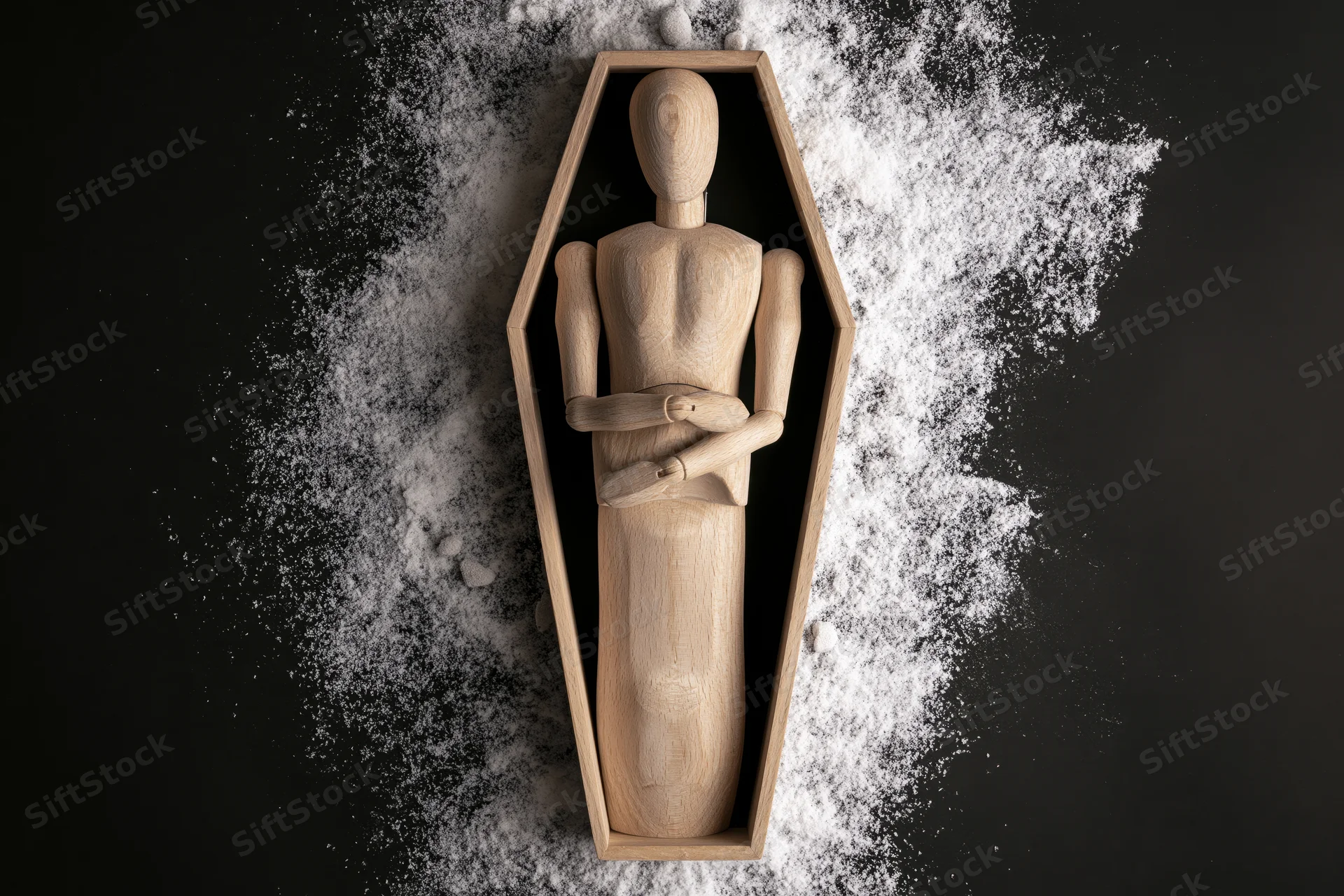 Wooden artist mannequin lying upright inside coffin-shaped frame on black surface with white powder scattered around