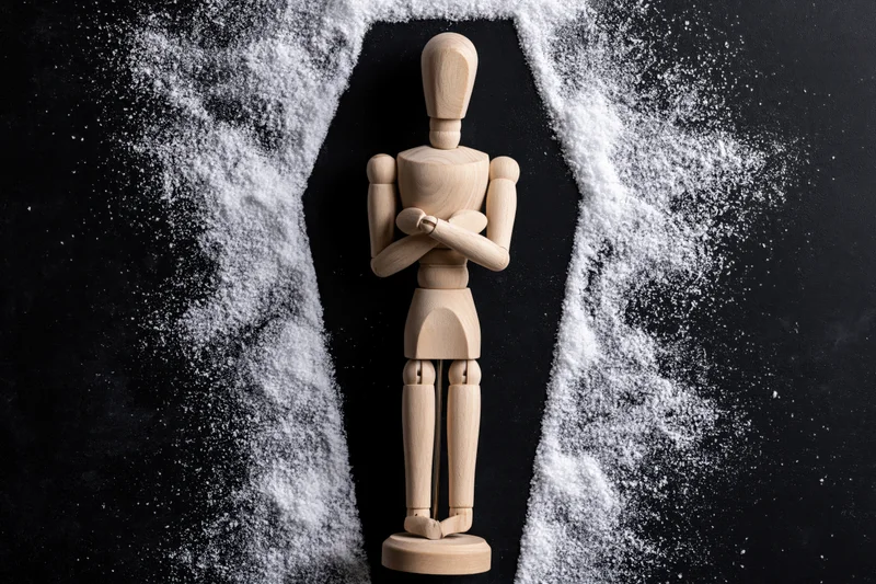 Wooden artist mannequin standing with arms crossed inside a white powder coffin shape on black background