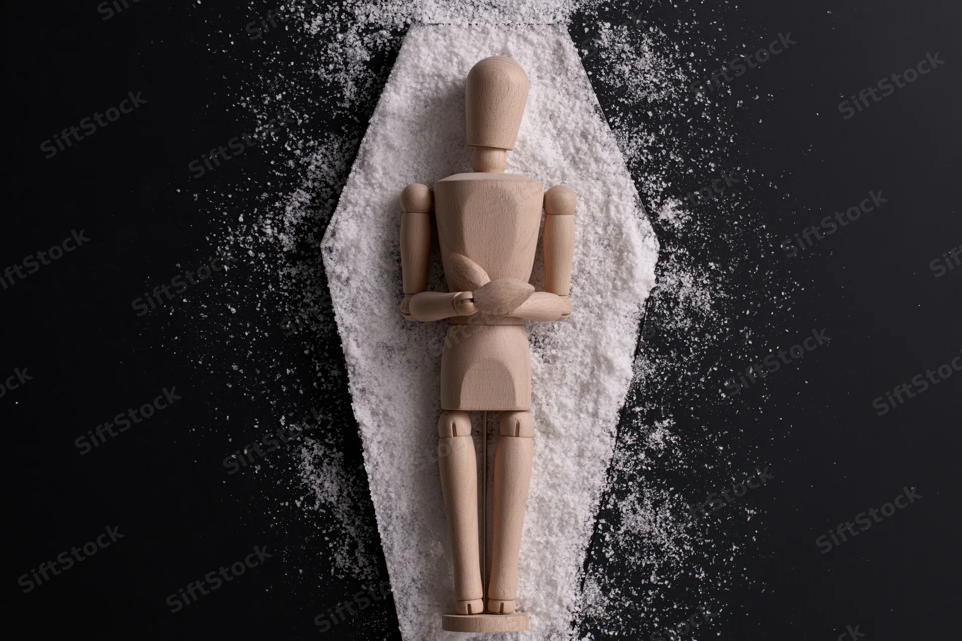 Wooden artist mannequin lying upright inside a coffin-shaped outline of white powder on black background
