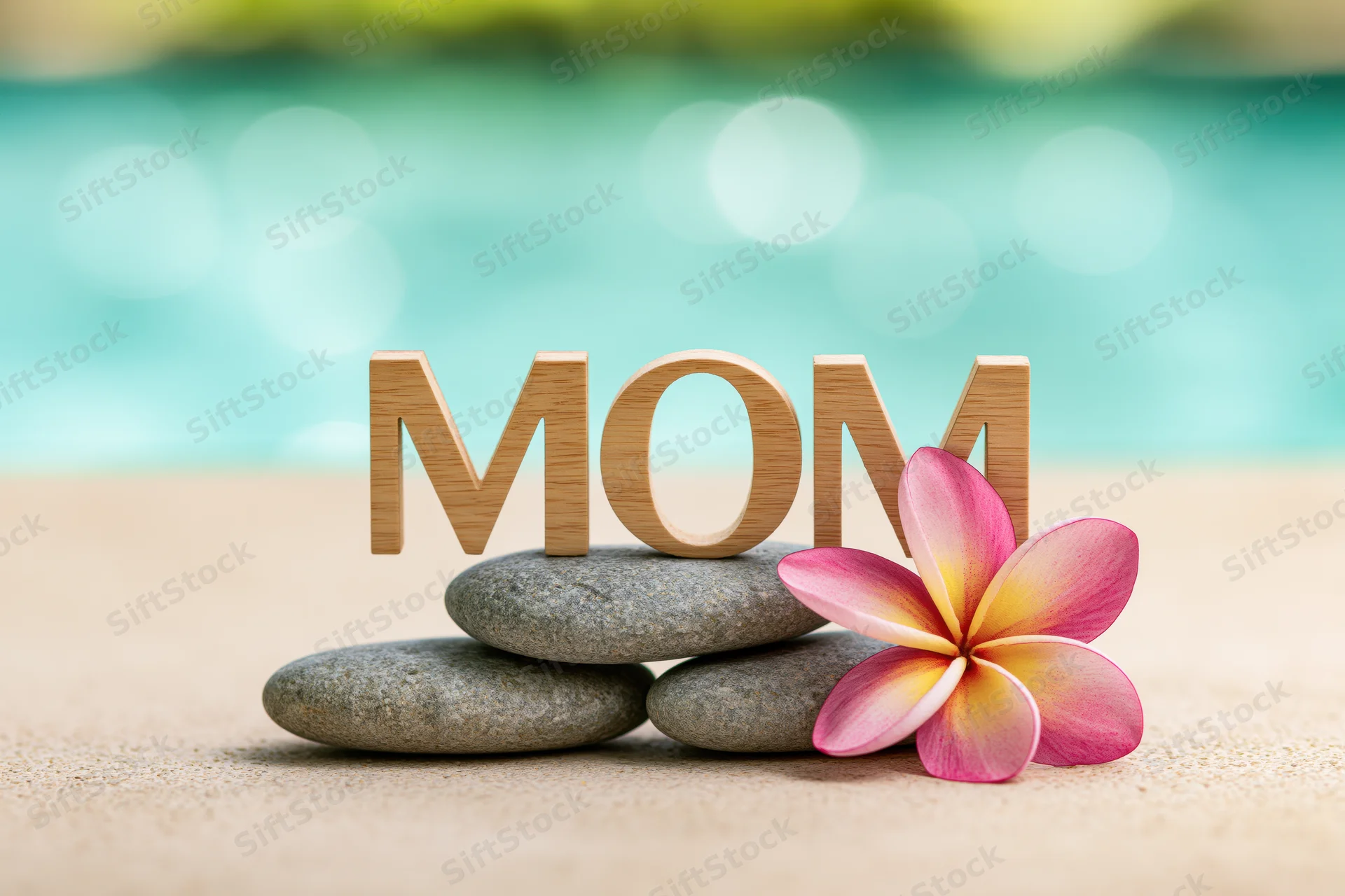 Wooden letters spelling mom on smooth stones with a frangipani flower on sandy beach near turquoise water
