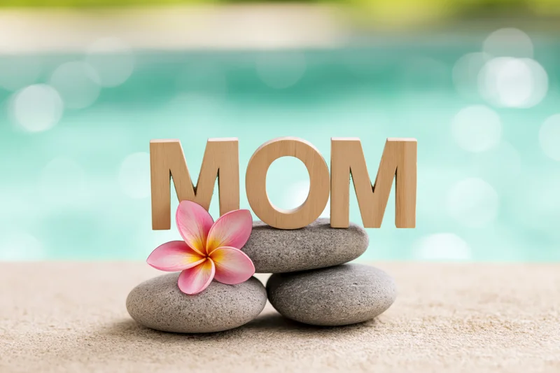 Wooden letters spelling mom on smooth stones with a frangipani flower by a turquoise poolside