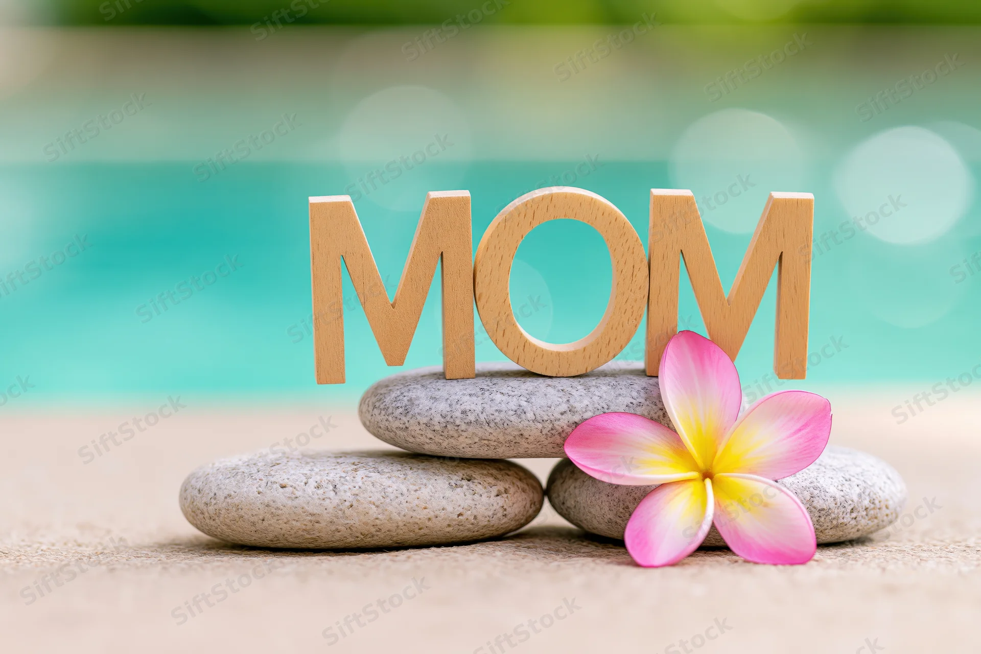Wooden letters spelling mom on smooth stones with a frangipani flower on sandy beach near turquoise water