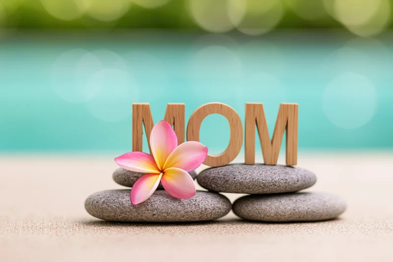 Wooden letters spelling mom on smooth stones with a frangipani flower, set against a blurred turquoise and green background