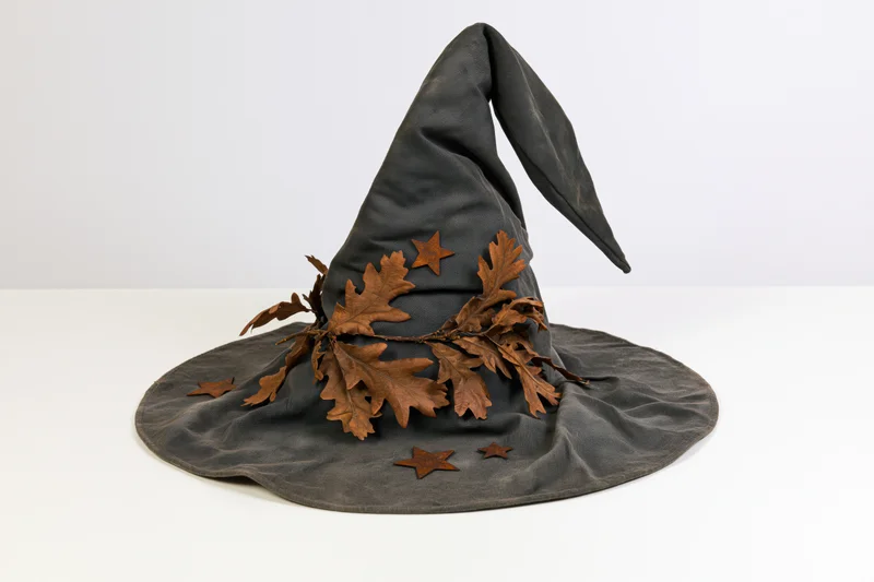 Dark grey witch hat with crooked brim adorned with dried oak leaves and stars on white surface