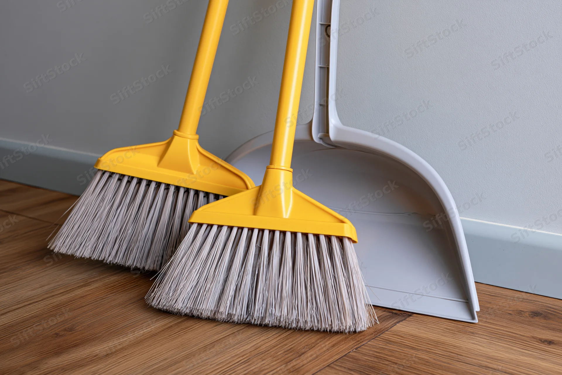 Two yellow-handled brooms and a grey dustpan leaning against a wall on a wooden floor