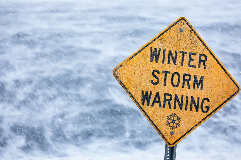 Yellow diamond-shaped road sign reading winter storm warning in falling snow